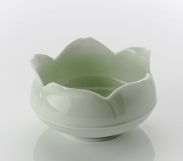 In front of me sits a small circular bowl that looks to be about 10cm in diameter and about 5cm deep. The bowl appears to be made from fine porcelain with thin walls that are only a couple of millimeters thick. The interior and exterior surface of the bowl is coated with a glossy almost transparent pale green glaze. The rim of the bowl blooms open into what looks like the outline of a flowering lotus. This is shaped by five identical triangular peaks that curve and fold out slightly from the rim of the bowl, each with a small concave indent that splits each peak into two. This is countered by negative space that cuts-in from the rim and forms an over extended V shape that organically curves like an opened trouser zip. This V shape is replicated between each flowering fold at five equal intervals around the perimeter of the bowl. This creates an undulated rhythm, giving the form its lotus effect. Looking at the bowl from the side, I see a protruding horizontal seam that divides the two hemispheres of the bowl. This makes the bottom half of the bowl appear to be tucked-in. If I pick up the bowl and Iook underneath, I can see that it rests on a fine foot rim about 5cm in diameter.