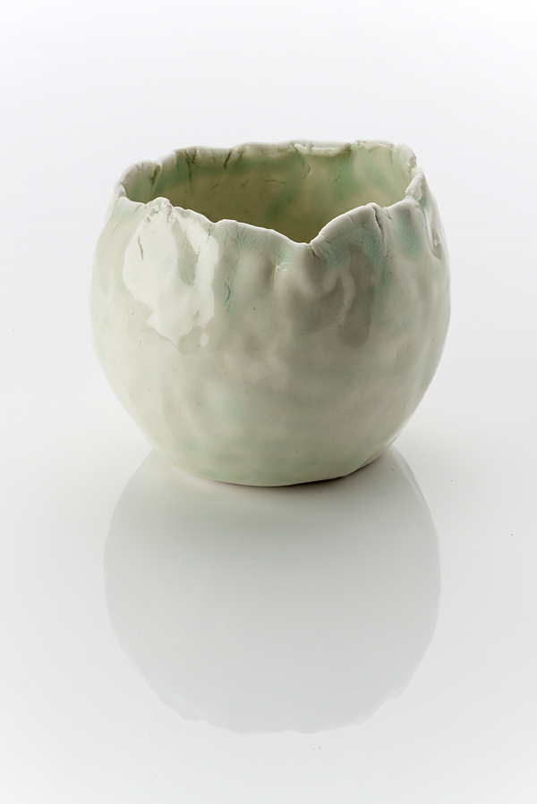 An oval shaped ceramic vessel made from a white clay body, washed over with a shiny glaze. The form is dimpled, roughly-made and unsteady with finger markings . The rim of the vessel looks like it has been pinched, which has stressed the clay to create slight vertical cracks.