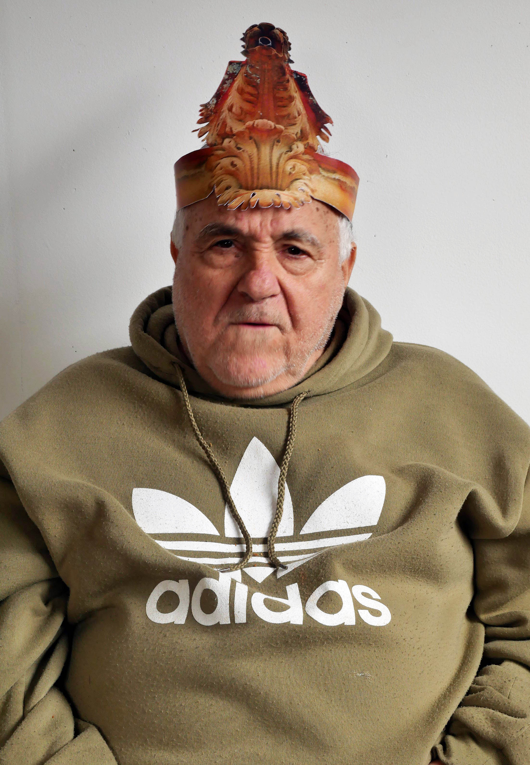 A portrait of old man looking straight at the camera. He has a grumpy expression on his face. He is wearing a paper party hat in the form of a Gargoyle. He is wearing a beige sports jumper with a large white Adidas logo printed on the front.