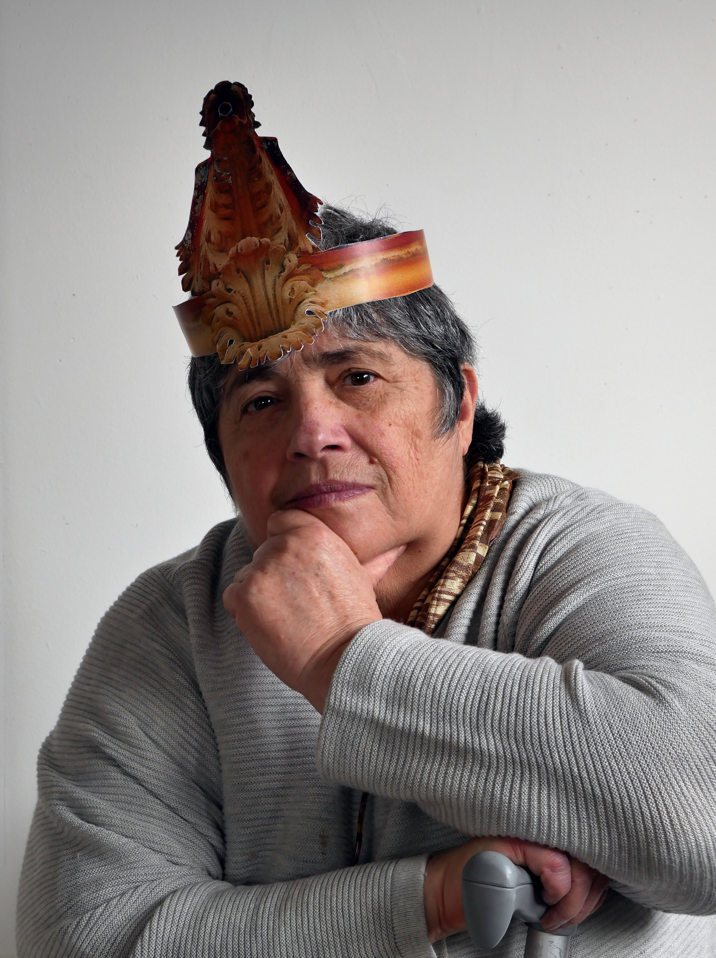 Portrait of a woman in her 70’s. She looks at the camera in a contemplative way; head tilted and hand on chin. On her head she wears a paper party hat in the form of a Gargoyle. She wears a pale grey jumper, which matches the handle of her walking stick on which she rests her folded arm.